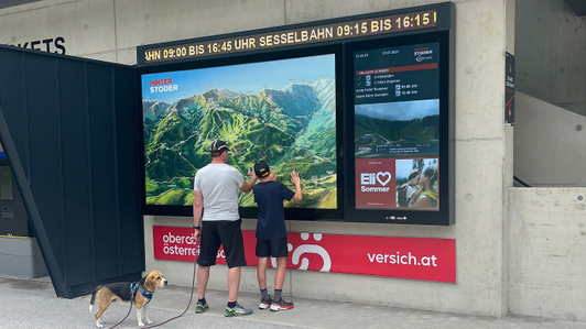 digitale Panorama-Infoanlage mit Laufschrift, Legendenbildschirm inkl. ELI-Spot und Werbeleiste mit OÖ Versicherung-Sujet mit Vater, Sohn und Hund davor, im Wandergebiet Hinterstoder