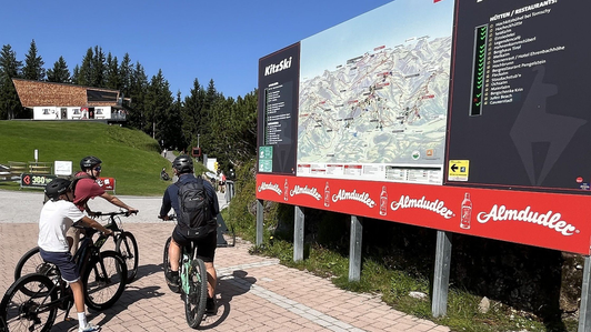 Panoramatafel mit LED-Legende und Almdudler-Werbeleiste, davor drei Mountainbiker, an der Bergstation der Hahnenkammbahn im Skigebiet KitzSki, Kitzbühel